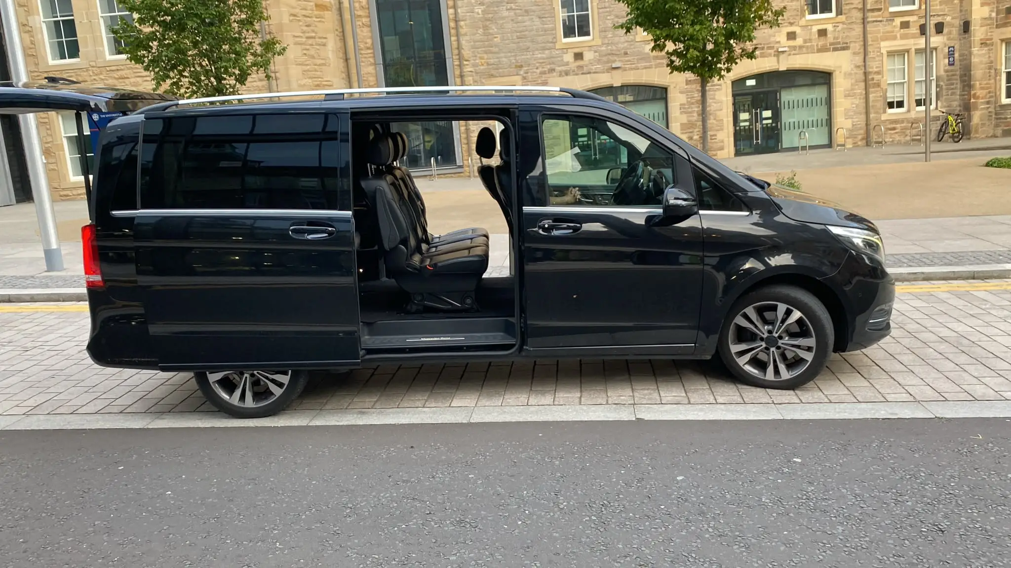 The black Mercedes V-Class side-on, sliding door open, outside a sandstone building